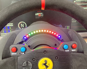 May include: A black racing steering wheel with a red stripe, a multi-colored LED display, and various buttons including "Mirror", "P-Lim", "Radio", and "Next". The steering wheel also features a Ferrari logo and a black suede rim.