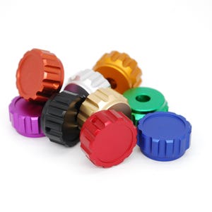 May include: A collection of colorful, machined metal caps in various colors, including red, orange, purple, black, gold, green, blue, and silver. The caps have a ridged, circular design and are arranged on a white background.