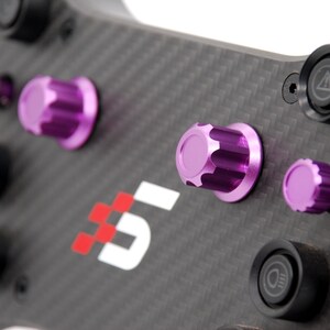 K&ouml;nnte beinhalten: Ein Rennlenkrad aus Kohlefaser mit strukturierter schwarzer Oberfl&auml;che, ausgestattet mit mehreren violett eloxierten Aluminium-Drehkn&ouml;pfen und schwarzen Drucktasten. In der Mitte befindet sich ein rot-wei&szlig;es Logo mit dem Text 51 sowie diverse Symbole.