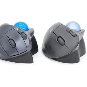 May include: Two ergonomic computer trackball mice. Each mouse is dark gray with a blue trackball and several buttons. The mice are designed for comfort and precision, with a curved shape and a built-in stand.