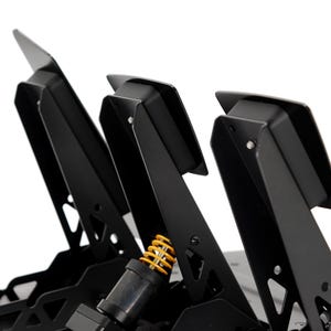 May include: Close-up of a black racing simulator pedal set. The set features three pedals with angled footplates. A yellow spring is visible. The pedals are mounted on a black base.