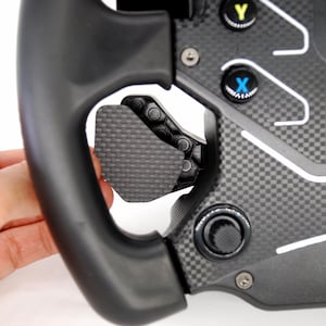 Può includere: Primo piano di un volante da corsa nero con dettagli in fibra di carbonio. Il volante presenta pulsanti con le etichette "Y" e "X" e una paletta strutturata. Il marchio "FANATEC" è visibile.