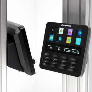 May include: Black Stream Deck control panel with a screen displaying app icons and adjustable knobs. The device is mounted on a silver metal frame, showcasing its sleek design and functionality for content creation and streaming.