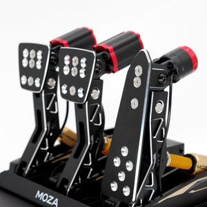 May include: Black and silver racing pedals with red detailing. The pedals feature a perforated design and are mounted on a black base. The brand name "MOZA" is visible on the base.