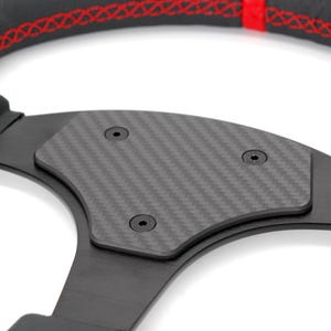Può includere: Primo piano di un volante nero con un inserto in fibra di carbonio e cuciture rosse. Il volante ha tre viti visibili.