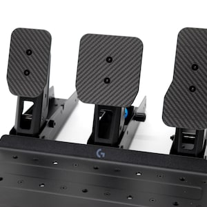 May include: Black racing simulator pedal set with three pedals. The pedals have a carbon fibre pattern on the foot pads and are mounted on a black metal base. The base has multiple mounting holes and the Logitech logo.