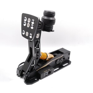 Puede incluir: Pedal de carreras negro con una placa de pie de metal y un resorte amarillo. El pedal está montado en una base negra con un acento dorado.