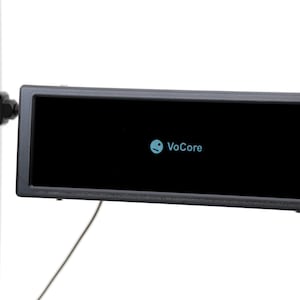 Puede incluir: Una pantalla de visualización rectangular negra con la palabra "VoCore" en azul claro. La pantalla está montada en un soporte negro fijado a un poste de metal plateado. Un cable negro se extiende desde la parte inferior de la pantalla.
