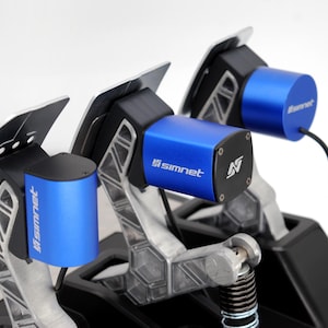 Puede incluir: Primer plano de un juego de pedales de simulador de carreras. El juego presenta pedales plateados con componentes negros y azules. Los componentes azules tienen el logotipo "simnet" y una "N" estilizada. Un cable negro se extiende desde uno de los cilindros azules.