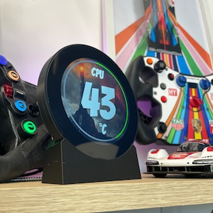 May include: A black circular device with a white display showing "CPU 43°C". The device is on a wooden surface with a toy car and a gaming controller in the background.