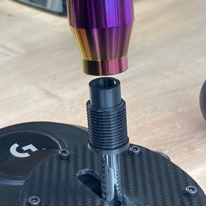 May include: A close-up of a black and silver shifter with a rainbow-colored shift knob. The shifter is attached to a black base with a carbon fiber pattern.