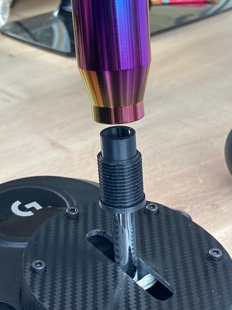 May include: A close-up of a black and silver shifter with a rainbow-colored shift knob. The shifter is attached to a black base with a carbon fiber pattern.