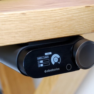以下が含まれることがあります： 木製のテーブルの下に取り付けられた、黒いSteelSeriesオーディオデバイス。大きなボリュームノブと、USB、48kHz/24bit、音量レベルを表示する小さなディスプレイがあります。ディスプレイには白い文字とグラフィックが表示されています。