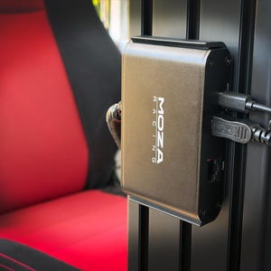 May include: A metallic grey "MOZA RACING" device is mounted on a black frame, with multiple cables plugged in. The device is rectangular with rounded edges. A red and black seat is visible in the background.