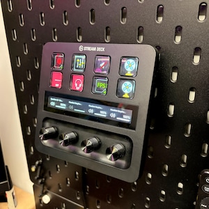 May include: Black Stream Deck device with multiple illuminated buttons and a small display, mounted on a black pegboard. The buttons feature icons for various functions. Below the display are four control knobs. A remote control is visible in the background.