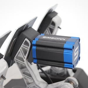 May include: A racing pedal set featuring a black and blue cylindrical motor housing with the text SIMSONN printed in white. The assembly includes metallic silver pedal arms with a structural lattice design and flat black pedal plates.