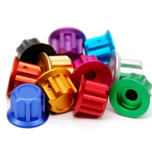 K&ouml;nnte beinhalten: Eine Ansammlung kleiner, zylindrischer Metallkn&ouml;pfe mit geriffeltem, zahnradartigem Au&szlig;endesign. Diese Komponenten weisen eine Vielzahl lebendiger, solider Farben auf, darunter Blau, Rot, Gr&uuml;n, Gold, Lila, Schwarz und Silber, jeweils mit einem Mittelloch.