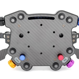 May include: A carbon fibre sim racing steering wheel hub featuring a central mounting plate, multiple black push buttons, and adjustable rotary dials in blue, red, orange, and purple. The unit includes side-mounted paddle shifters and labels for ABS and TC.