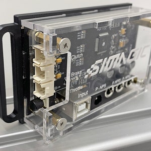 Puede incluir: Una caja de plástico transparente con un asa negra, que contiene una placa de circuito impreso con varios conectores y etiquetas, incluyendo "Throttle", "Brake", "Clutch" e "Input". La caja tiene la palabra "SIMAGIC" impresa en ella.
