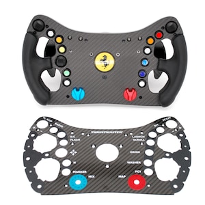 Può includere: Due volanti neri e grigi per simulatore di corse. Il volante superiore ha un logo Ferrari e diversi pulsanti colorati. Il volante inferiore ha il logo Thrustmaster e vari pulsanti etichettati. Entrambi su sfondo bianco.