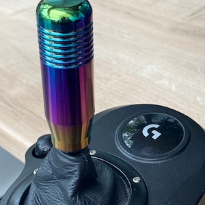 May include: A black racing wheel with a leather gear shifter and a rainbow colored metal shifter knob. The wheel has a G logo on the front.