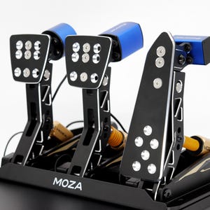 May include: A black and silver racing simulator pedal set with three pedals. The pedals have silver accents and are mounted on a black base. The top of the pedals are blue. The brand name "MOZA" is visible on the base.