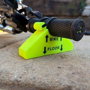 May include: A neon yellow plastic bicycle handlebar support block featuring a hook for the handlebar grip. The block is printed with the text BIKE above an upward arrow and FLOOR below a downward arrow to indicate the correct orientation for use.