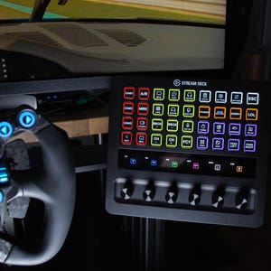 Peut inclure: Un panneau de contrôle Stream Deck noir avec plusieurs boutons éclairés et une rangée de boutons. Le panneau est positionné à côté d'un volant de course avec des boutons bleus et rouges. Un écran d'ordinateur est visible en arrière-plan.