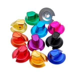 Può includere: Una collezione di undici manopole di controllo circolari in vari colori metallizzati, tra cui verde, giallo, arancione, rosso, azzurro, viola, nero, rosa e blu scuro, ciascuna dotata di un'impugnatura centrale rialzata e un piccolo foro laterale.