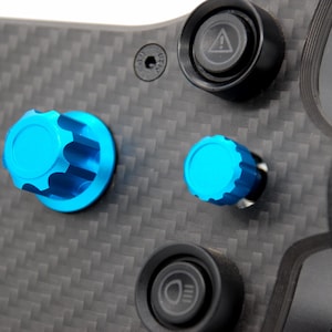 K&ouml;nnte beinhalten: Ein Lenkradpanel aus Kohlefaser mit zwei hellblauen, ger&auml;ndelten Drehkn&ouml;pfen, einem schwarzen Druckknopf mit wei&szlig;em Warndreieck-Symbol und einem schwarzen Druckknopf mit wei&szlig;em Scheinwerfer-Symbol auf einer texturierten, dunkelgrauen Oberfl&auml;che.