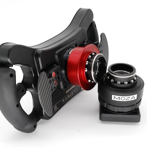 May include: A black racing steering wheel with a red and black quick release system. The quick release system is detached from the steering wheel and is labelled "MOZA".