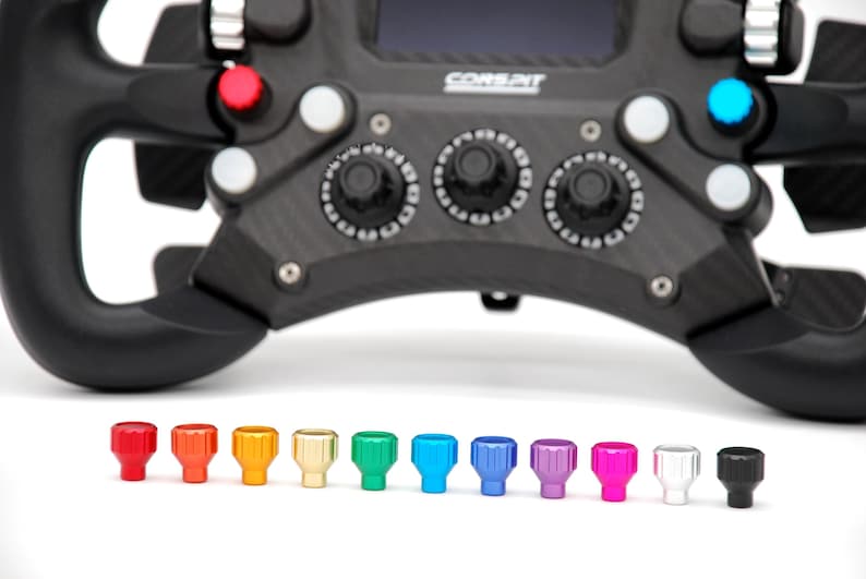 Peut inclure: Un volant de course noir avec le texte CORSPIT, dot&eacute; de plusieurs boutons et cadrans rotatifs. Sous le volant se trouvent onze petits boutons cylindriques stri&eacute;s dans un spectre de couleurs incluant rouge, orange, jaune, or, vert, bleu, violet, rose, blanc et noir.