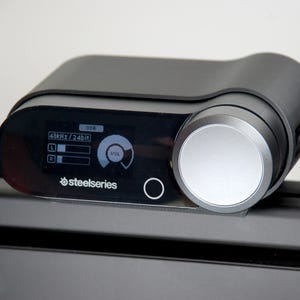 以下が含まれることがあります： 黒い持ち手と「But First, Coffee」のテキストが黒い太字でプリントされた白いセラミックマグ。マグは木製の表面に置かれています。マグの高さは約10cmです。