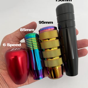 May include: Four different gear shift knobs in various colors and sizes. The knobs are made of metal and have a textured surface. The knobs are labeled with their measurements in millimeters: 85mm, 95mm, and 130mm. One knob is red and says "6 Speed".