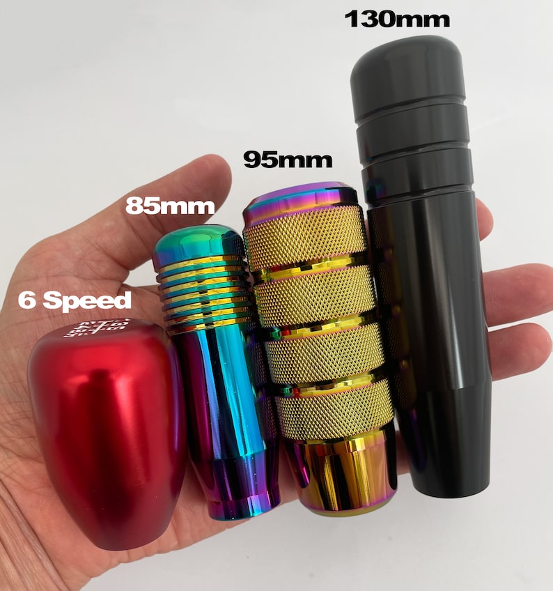 May include: Four different gear shift knobs in various colors and sizes. The knobs are made of metal and have a textured surface. The knobs are labeled with their measurements in millimeters: 85mm, 95mm, and 130mm. One knob is red and says "6 Speed".