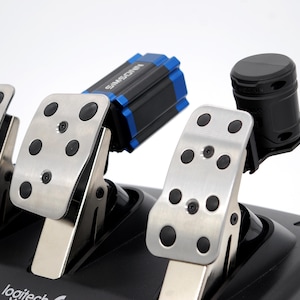 Puede incluir: Juego de pedales para volante de juego con pedales plateados y detalles en negro. Los pedales tienen agarres de goma negra y están montados sobre una base negra. También se ve un dispositivo negro y azul.