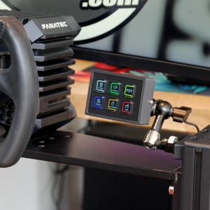 May include: A black racing simulator setup featuring a Fanatec steering wheel and a control panel with illuminated buttons. The wheel has carbon fiber accents and multiple buttons. The control panel has six buttons with various icons.
