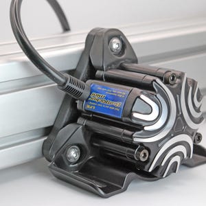 Puede incluir: Un actuador lineal eléctrico negro con una tapa final de patrón plateado y negro. El actuador está montado en una vía plateada y tiene una etiqueta azul que dice "Brackit Mini".