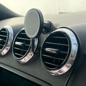 Puede incluir: Interior de coche con tres salidas de aire circulares con molduras plateadas y un soporte magnético negro para teléfono. El salpicadero es de color gris oscuro y las rejillas de ventilación tienen lamas ajustables. El soporte del teléfono está fijado al salpicadero.