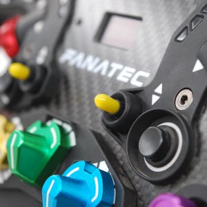May include: A close-up shot of a racing wheel, branded with FANATEC. The wheel has a carbon fibre texture and features an array of coloured buttons and knobs, including red, yellow, green, blue, and purple.