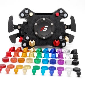Op de afbeelding: Een zwart en koolstofvezel racestuur met diverse gekleurde knoppen en wijzerplaten. Het stuur heeft een centrale naaf met het nummer "5" in rood en wit. Een assortiment gekleurde knoppen bevindt zich onder het stuur.
