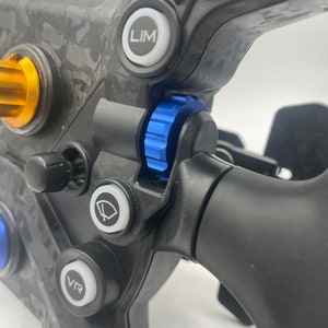 May include: Close-up of a black racing steering wheel with a blue and silver knob, a white button labelled "VR", and a white button labelled "LIM".
