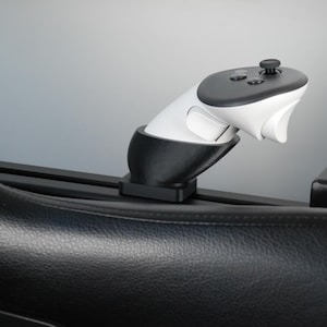 Puede incluir: Un mando de videojuegos blanco y negro montado en un soporte negro. El mando tiene un joystick y varios botones. El soporte está fijado a una superficie negra, posiblemente un asiento o una consola. El fondo es gris claro.