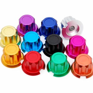 May include: A collection of eleven cylindrical knobs with fluted sides and flat tops, each featuring a distinct metallic finish in colours including red, blue, green, yellow, orange, purple, pink, black, and silver, arranged in a cluster.