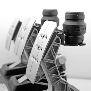 Könnte beinhalten: Ein Satz von drei schwarzen und silbernen Rennsportpedalen mit einer strukturierten Oberfläche. Die Pedale sind mit einem Metallrahmen verbunden und haben eine schwarze Basis. Die Pedale sind mit dem Wort "STRANGE" beschriftet.