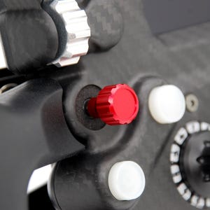 Peut inclure: Un composant de volant en fibre de carbone noire dot&eacute; d'un bouton cylindrique rouge pro&eacute;minent, de deux boutons circulaires blancs et d'une molette noire avec des marquages num&eacute;riques blancs. L'ensemble inclut une bague de r&eacute;glage m&eacute;tallique.