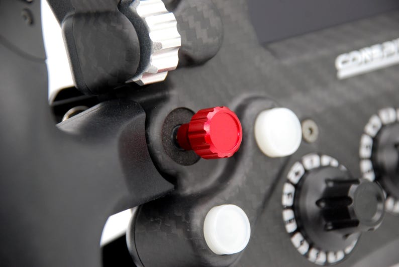 Peut inclure: Un composant de volant en fibre de carbone noire dot&eacute; d'un bouton cylindrique rouge pro&eacute;minent, de deux boutons circulaires blancs et d'une molette noire avec des marquages num&eacute;riques blancs. L'ensemble inclut une bague de r&eacute;glage m&eacute;tallique.