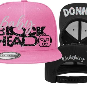 May include: Pink snapback hat with the text "Baby Big Head" in silver glitter lettering and a robot graphic. A black snapback hat with the name "Donnie" in silver glitter and the name "Wahlberg" in white script is also shown.