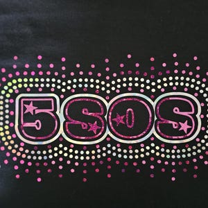 May include: Black graphic tee featuring the text "5SOS" in a pink, outlined font with star accents. The design is surrounded by a border of pink and white dots, with a gradient of colors.