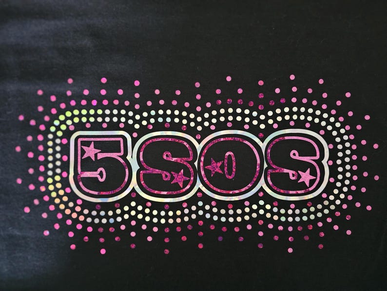 May include: Black graphic tee featuring the text "5SOS" in a pink, outlined font with star accents. The design is surrounded by a border of pink and white dots, with a gradient of colors.